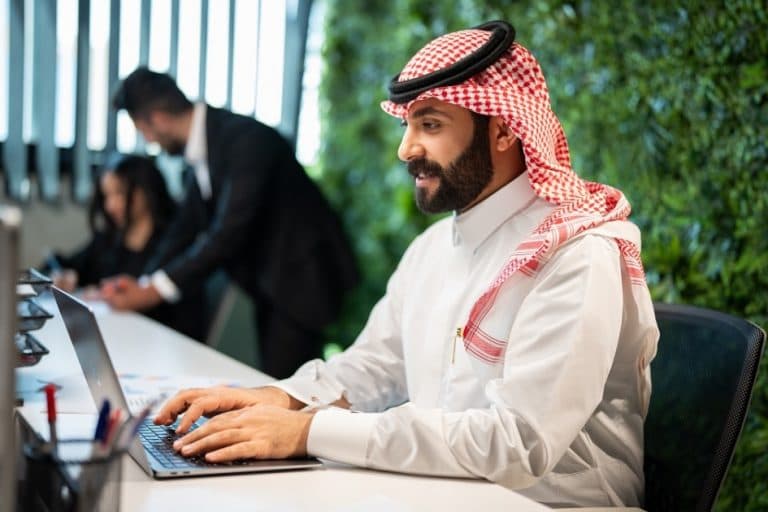 رجل سعودي يرتدي الثوب والغترة يعمل على جهاز الكمبيوتر، يرمز إلى المهنية والخبرة في إعداد دراسات الجدوى الاقتصادية